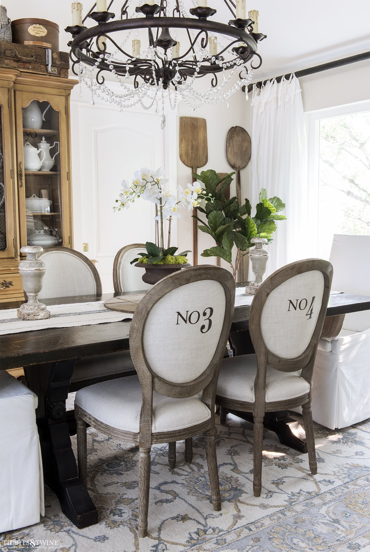 round french linen chairs with numbers on the back in dining room with white orchid on the table and vintage bread paddles hanging on the wall