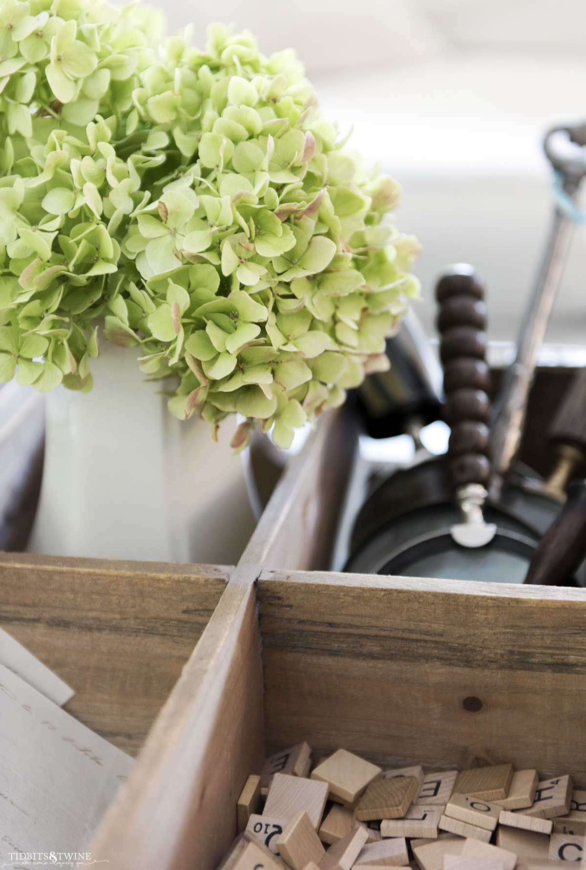 white ironstone vase filled with green hydrangea that are drying in a wooden box with magnifying glasses and scrabble tiles on coffee table