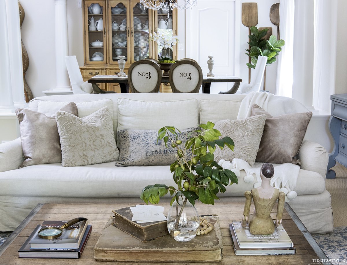 linen slipcovered sofa with beige decorative pillows and industrial coffee table in front and dining room in background with french round numbered chairs