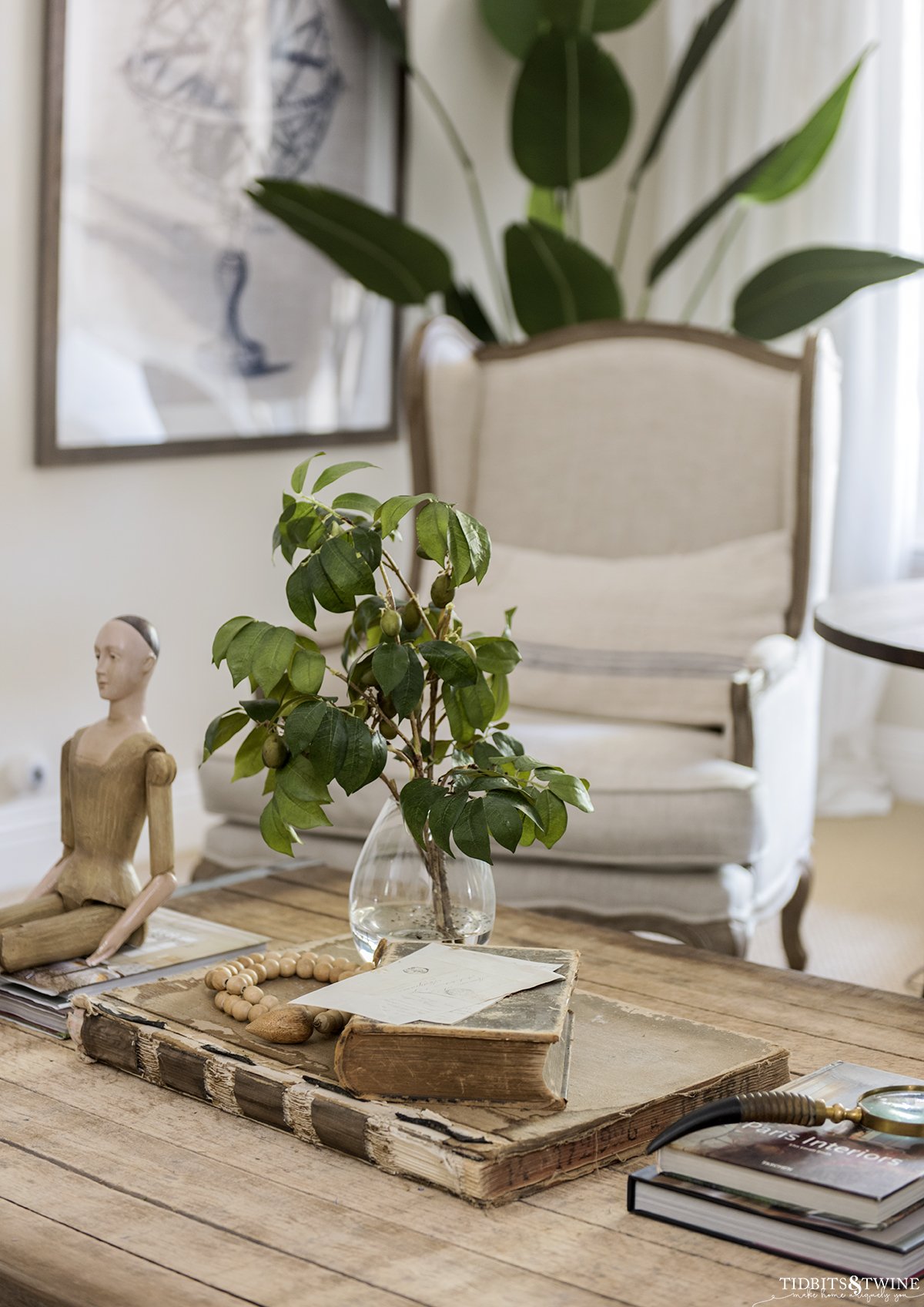 neutral living room with wooden top coffee table with old books on top and a vase with olive branches next to santos doll with french wingback chair in background