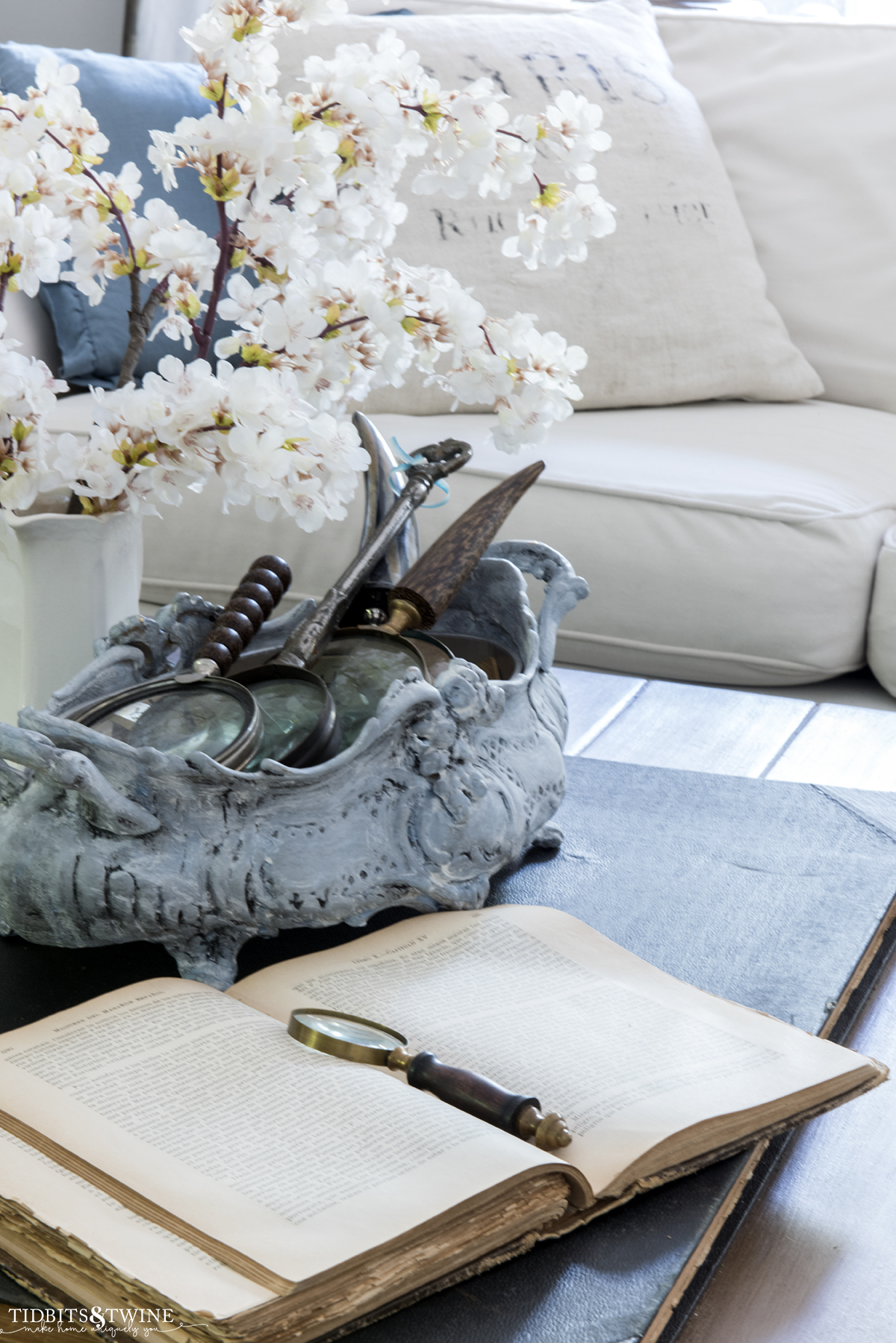 cherry blossom stems in an ironstone pitcher on coffee table with an antique book and sectional sofa in background with french pillows