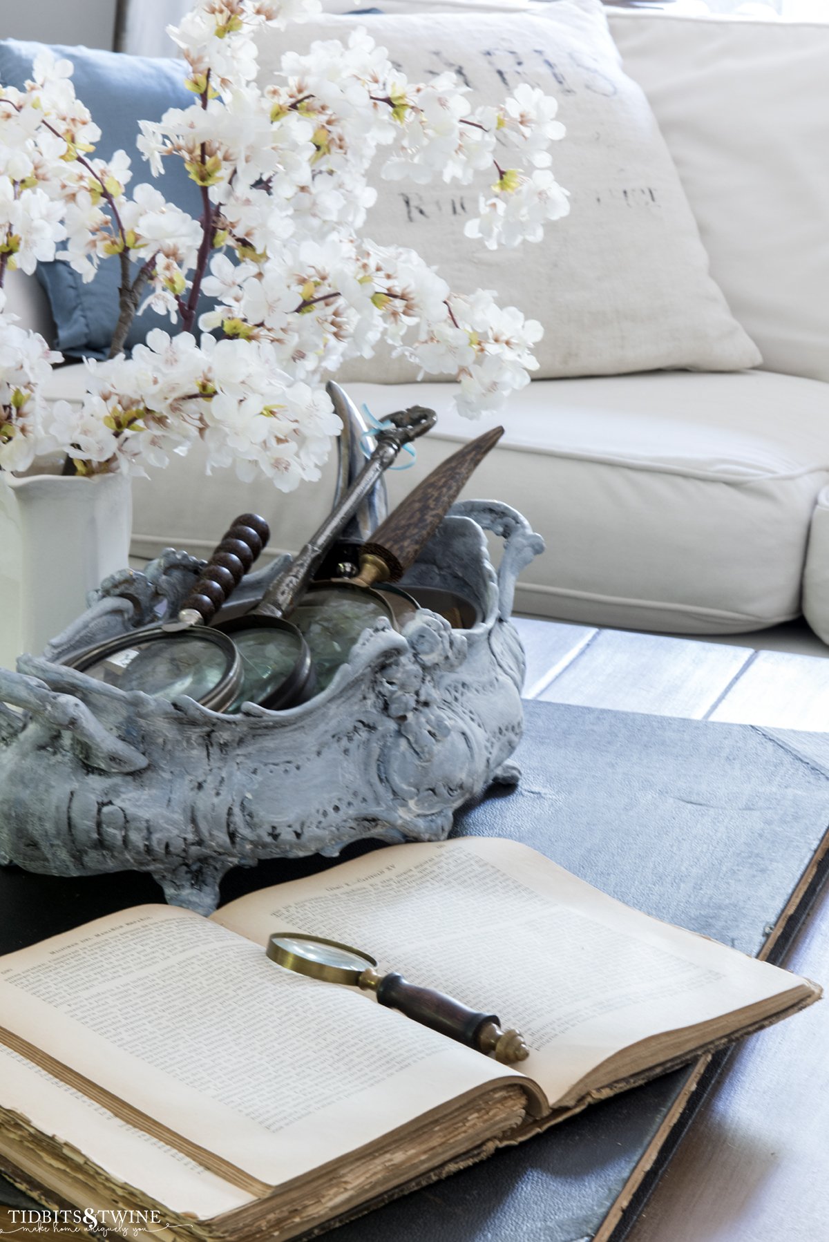 cherry blossom stems in an ironstone pitcher on coffee table with an antique book and sectional sofa in background with french pillows