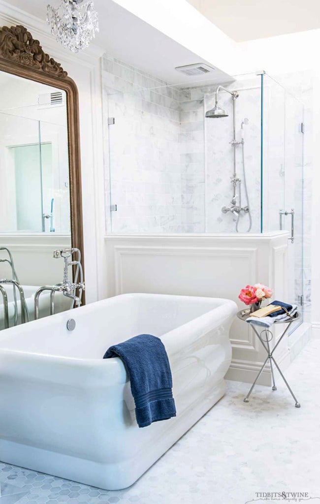 white marble bathroom with freestanding tub next to pony wall and large wood french mirror behind the tub