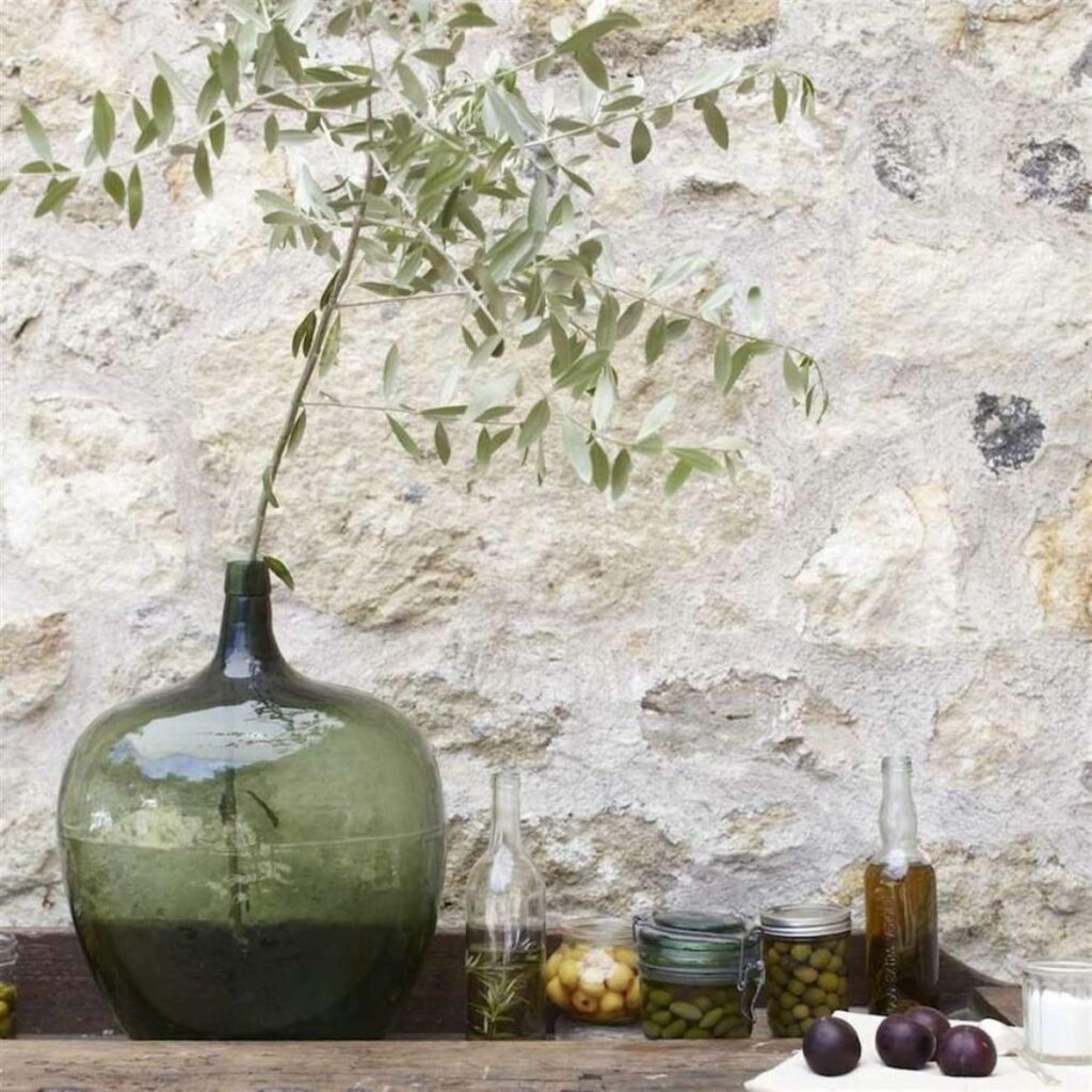 antique green demijohn holding a single olive branch next to jars full of olives and olive oil against a rock wall