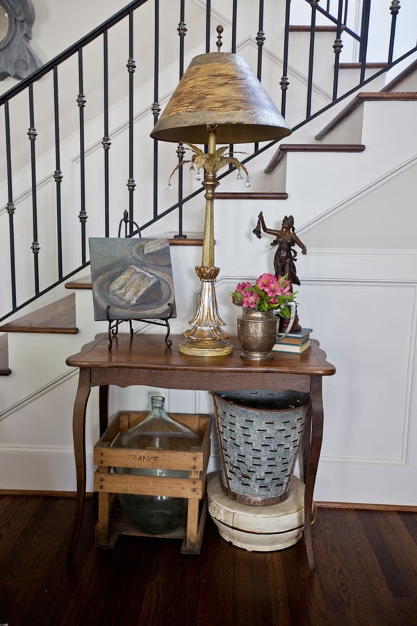 small table against a stair wall holding a lamp and a silver pitcher with flowers and underneath an antique olive bucket and crated demijohn