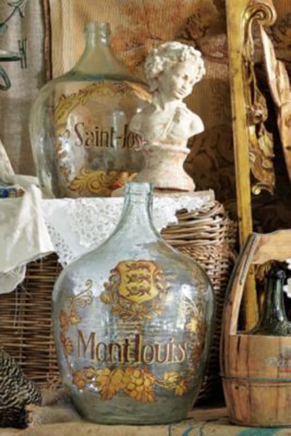 closeup of antique wine demijohn with gold paint on a clear bottle and basket and bust in the background