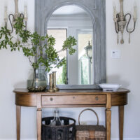 trumeau mirror above entry table flanked by french sconces with demijohn and basket underneath the table