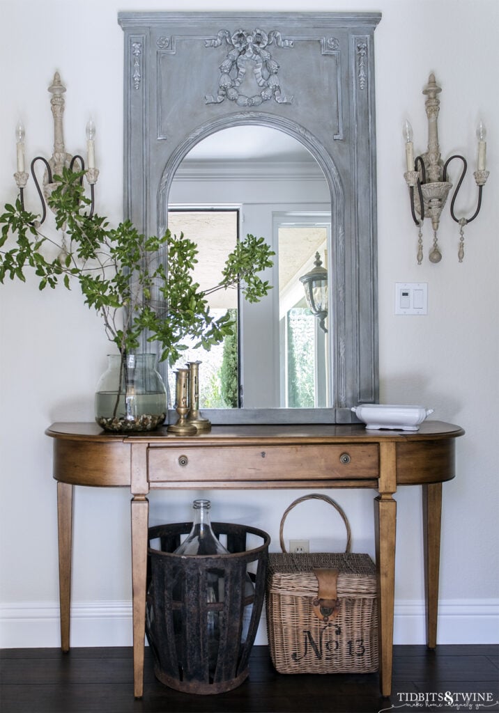 trumeau mirror above entry table flanked by french sconces with demijohn and basket underneath the table