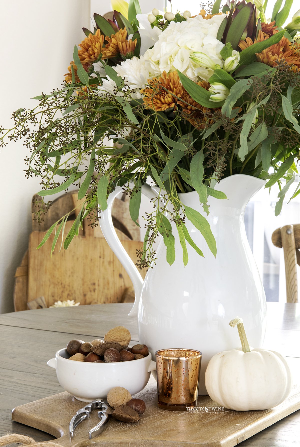 fall flower arrangement with white hydrangea and rust chrysanthemum in white pitcher on kitchen table with bowl of nuts