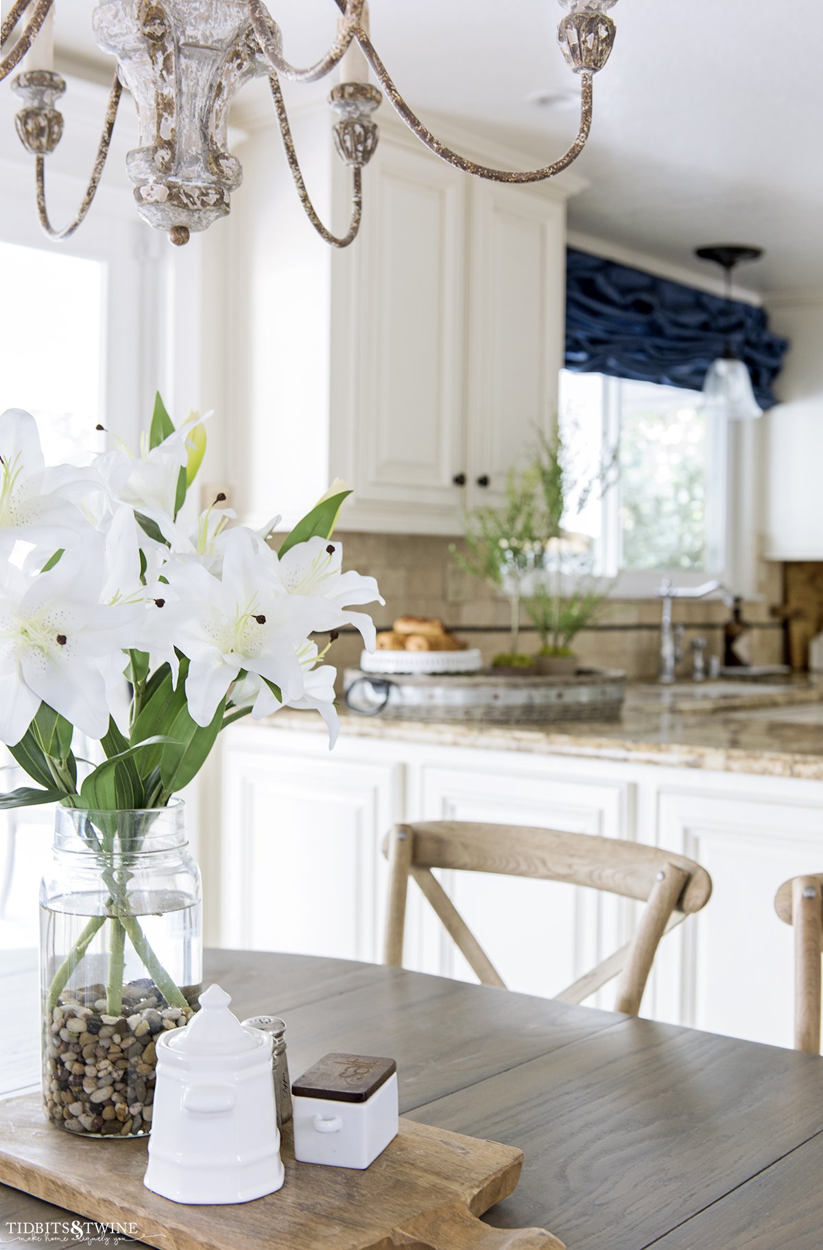 farmhouse kitchen with white lillies in center of table crossback chairs and myrtle topiaries in background