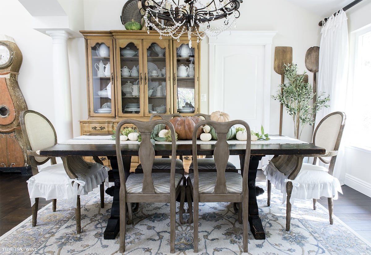 french dining room decorated for fall with pumpkins down center of table chairs with ruffle seat covers and hutch with white ironstone