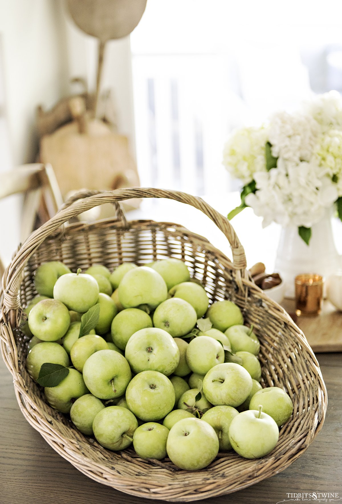 harvest basket full of green apples on kitchen table with white hydrangeas in a pitcher in the background with a collection of breadboards in the corner