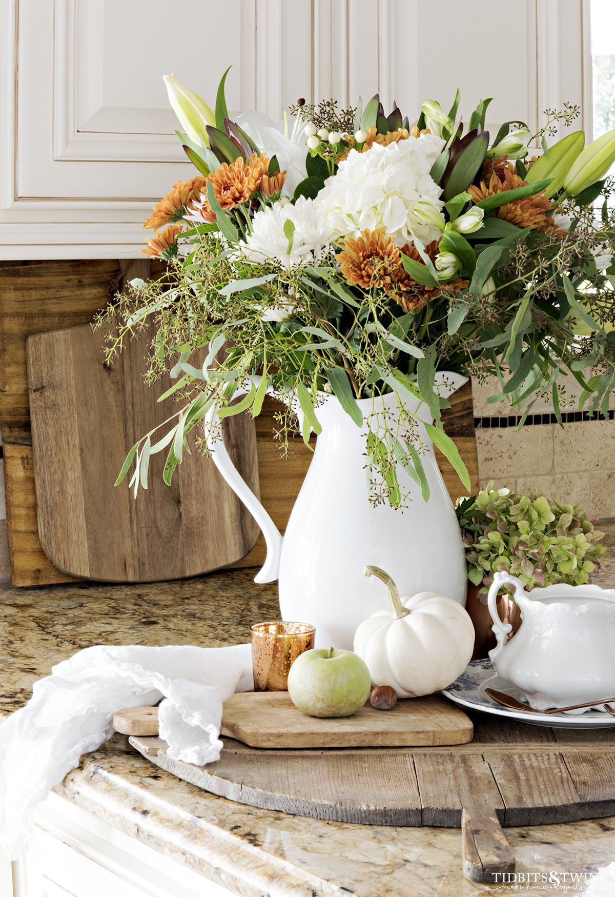 kitchen counter with fall vignette of white green and copper flowers in white pitcher on antique bread board with white pumpkins and apples