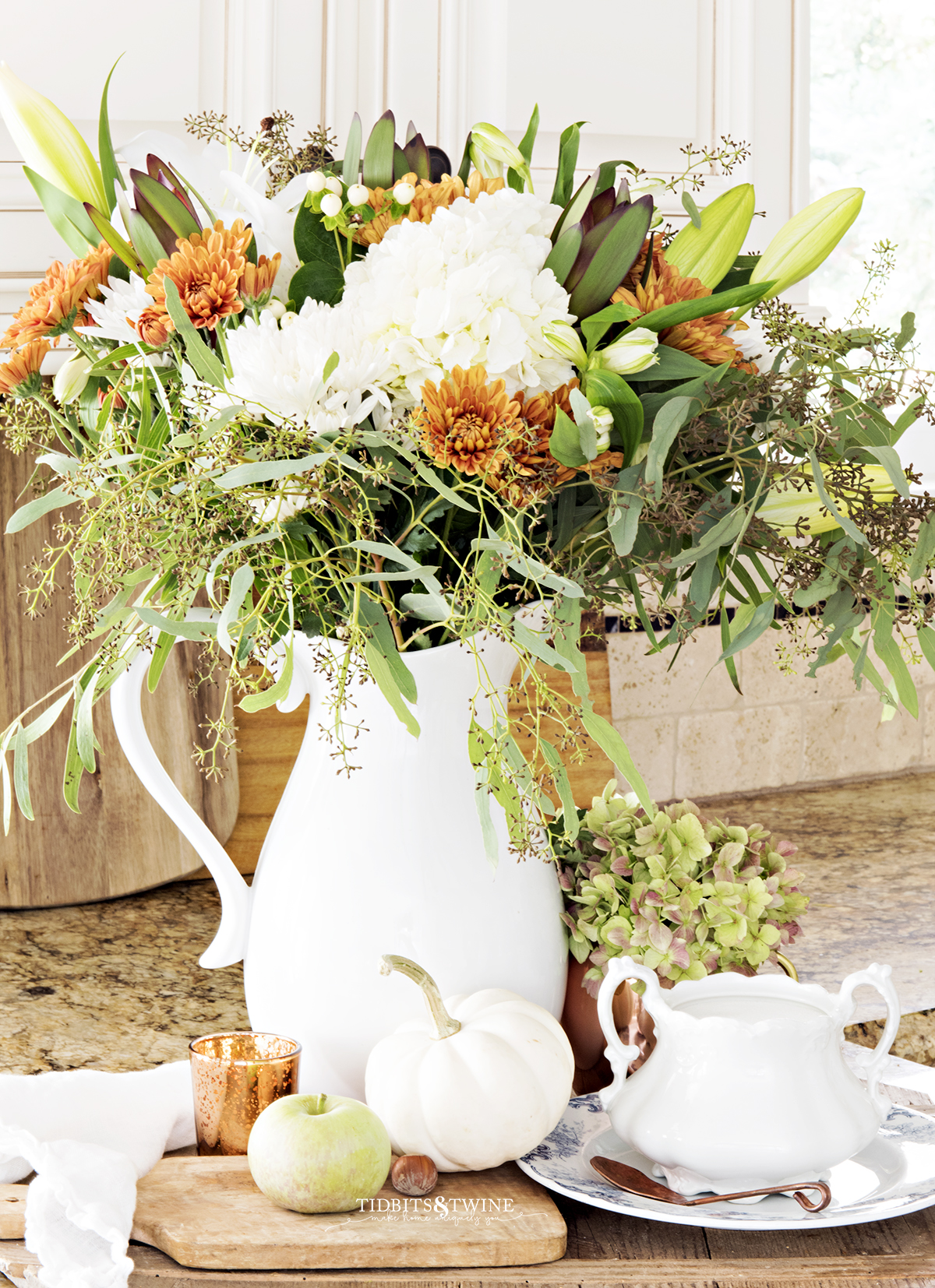 kitchen counter with fall vignette of white green and copper flowers in white pitcher on antique bread board with white pumpkins and apples