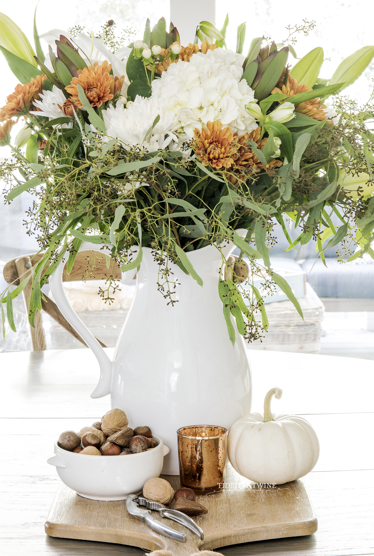 fall flower arrangement with white hydrangea and rust chrysanthemum in white pitcher on kitchen table with bowl of nuts