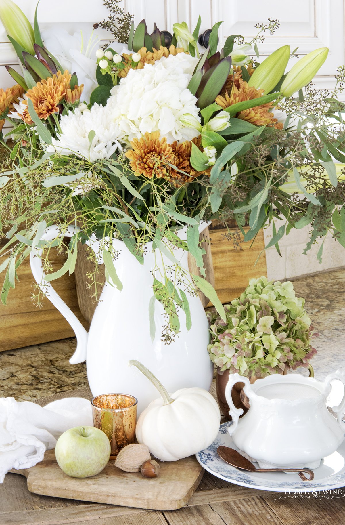 kitchen counter with fall vignette of white green and copper flowers in white pitcher on antique bread board with white pumpkins and apples