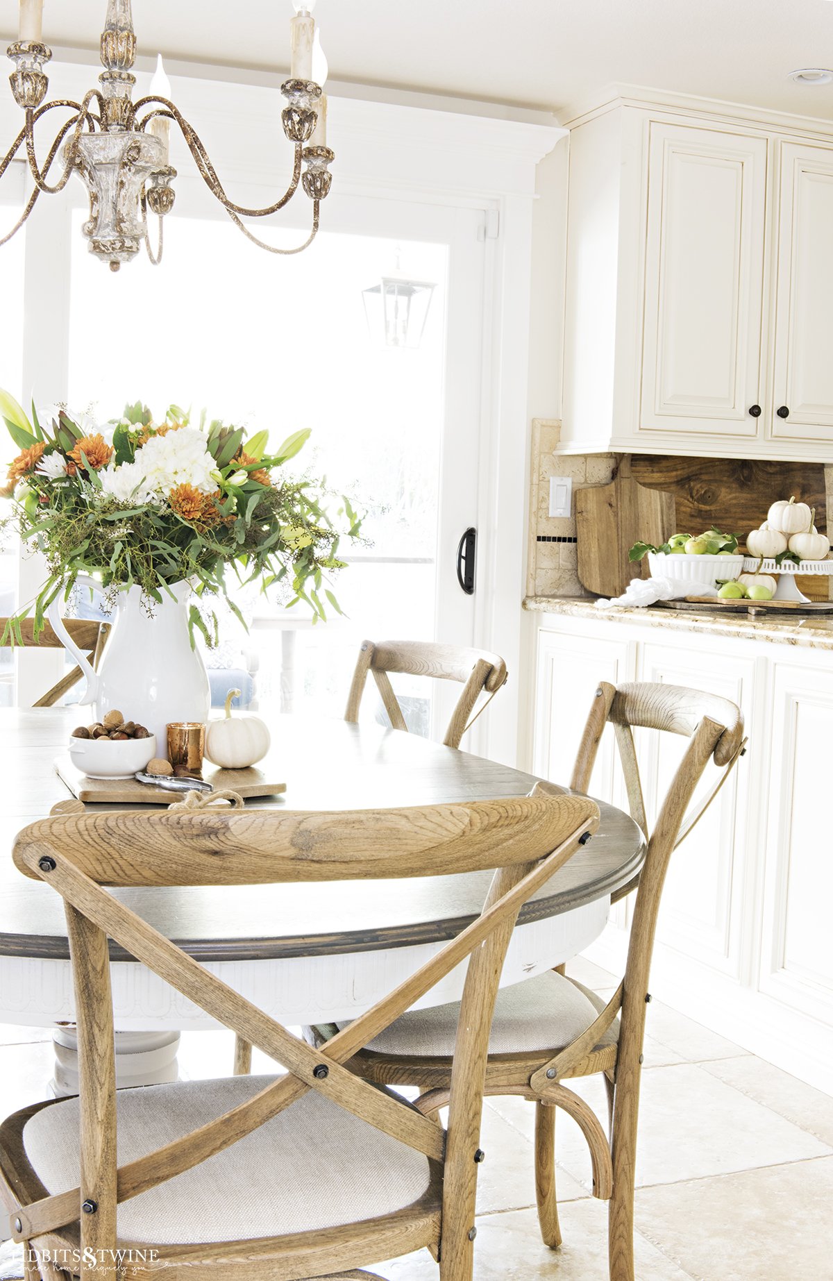 french country fall kitchen decor with table with white pitcher of fall flowers and crossback chairs and bowl of apples and pumpkins on countertop