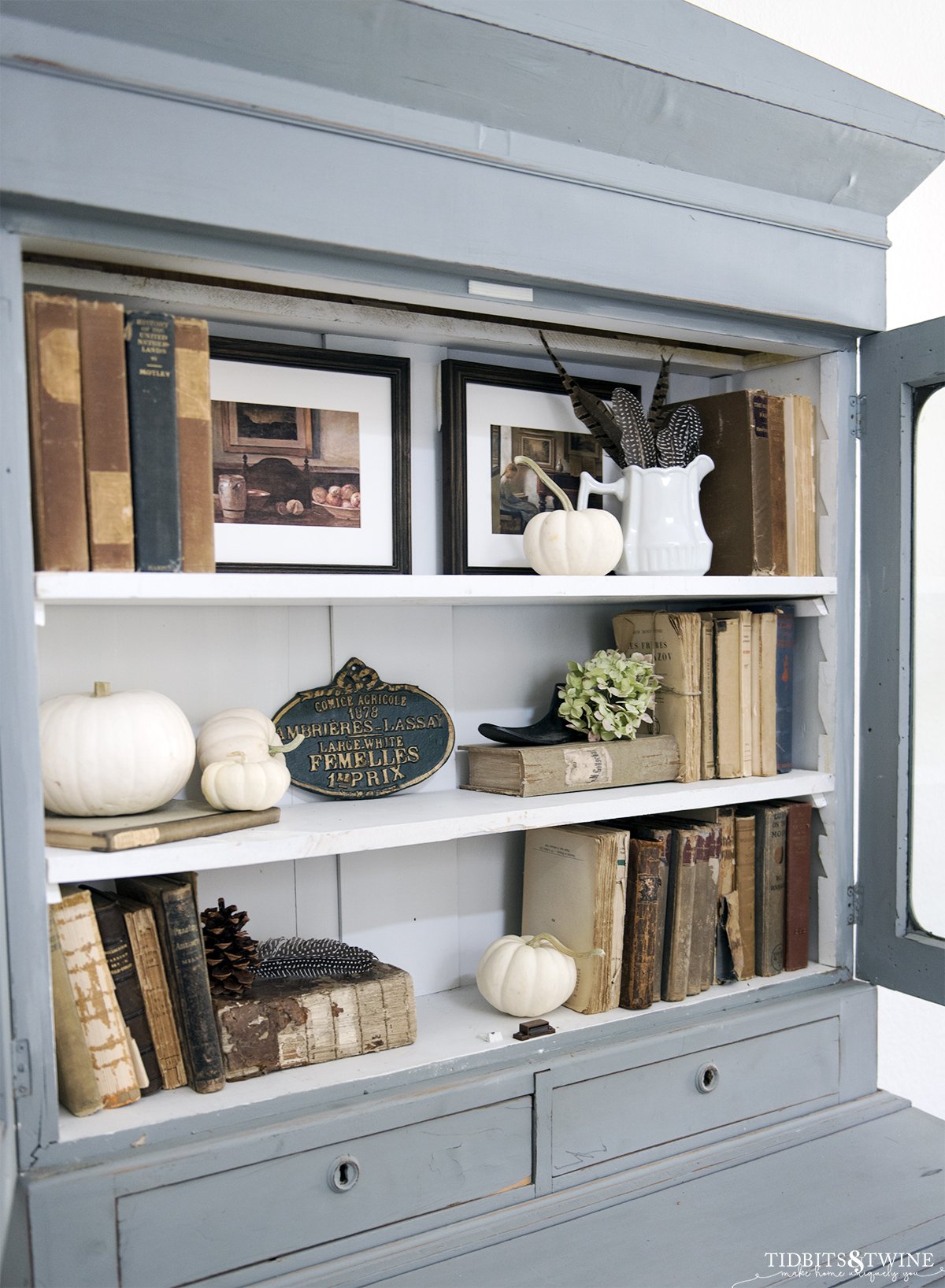 antique blue cabinet with three white shelves styled with old books art pumpkins and hydrangea for fall