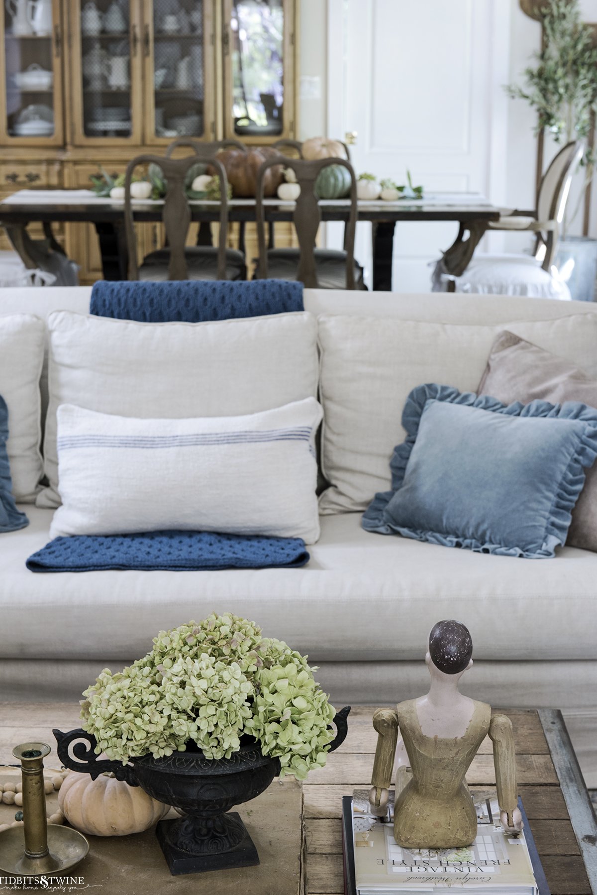 coffee table decorated for fall in front of slipcovered linen sofa with blue throw and dining room in background