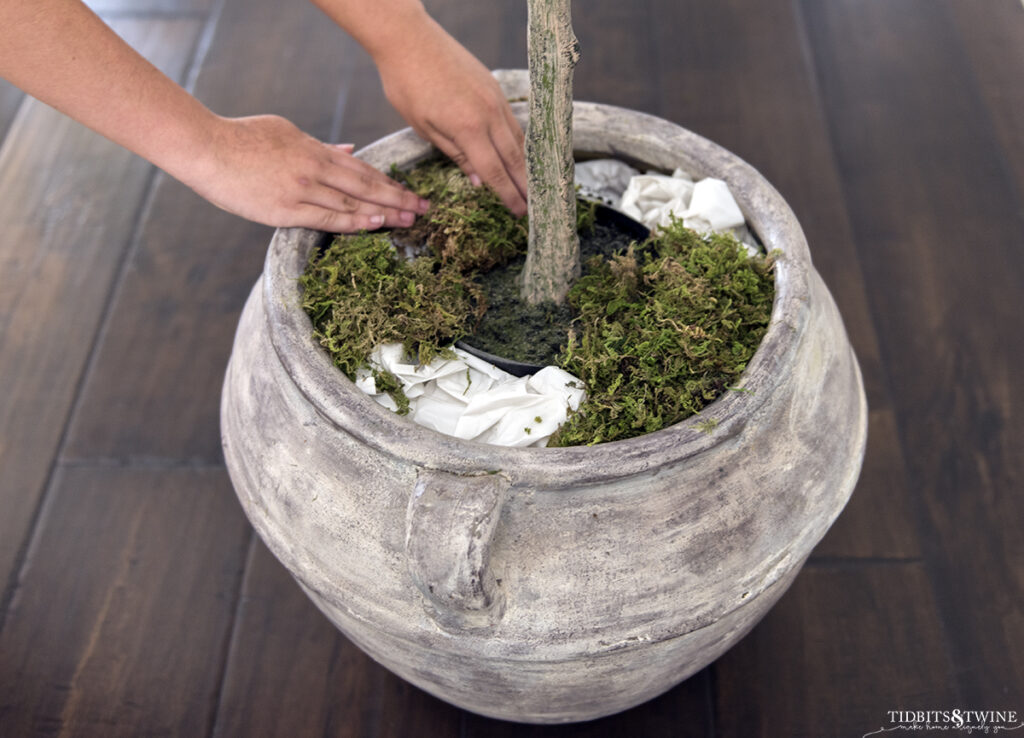 brown clay pot with artificial plant surrounded by stuffed grocery bags and moss being added on top