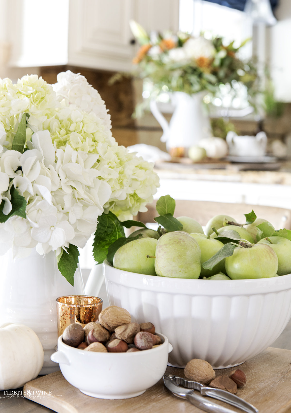 bowl of green apples next to bowl of nuts and white pitcher of white hydrangeas on kitchen table with fall flowers in background