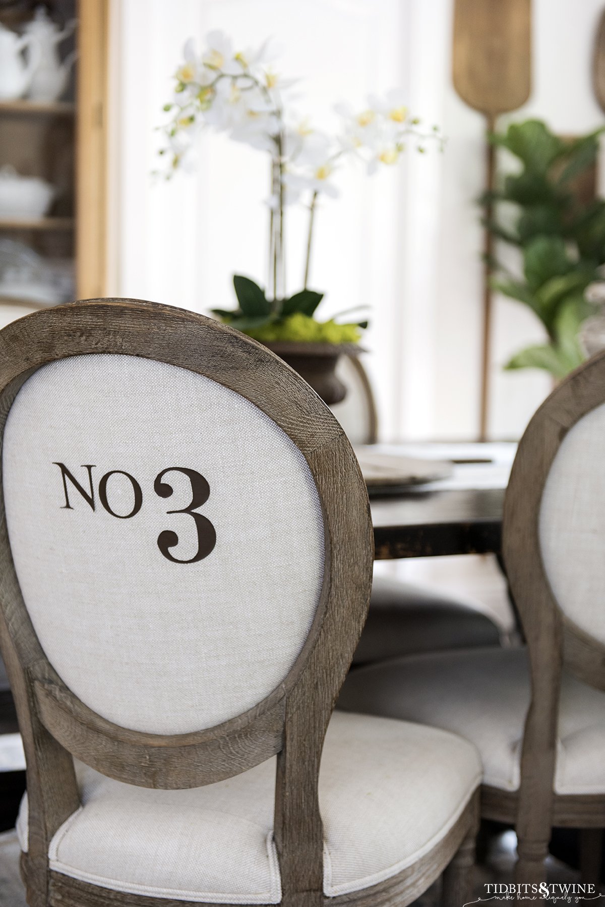 French round back chair with beige linen upholstery with numbers on the back in a dining room with white orchid on table