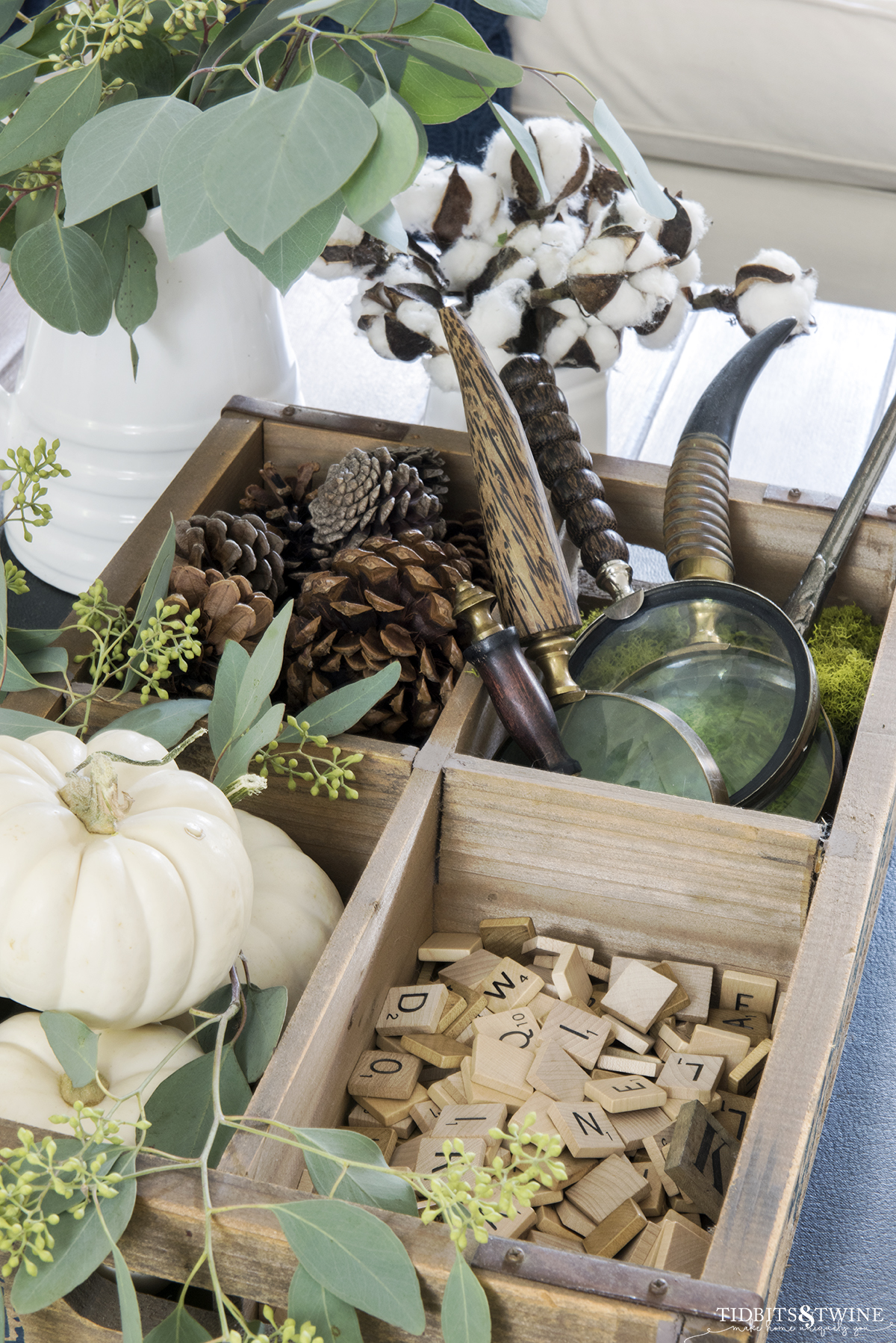 wooden crate with four cubbies one with white pumpkins and eucalyptus one magnifying glasses one pinecones and one scrabble tiles