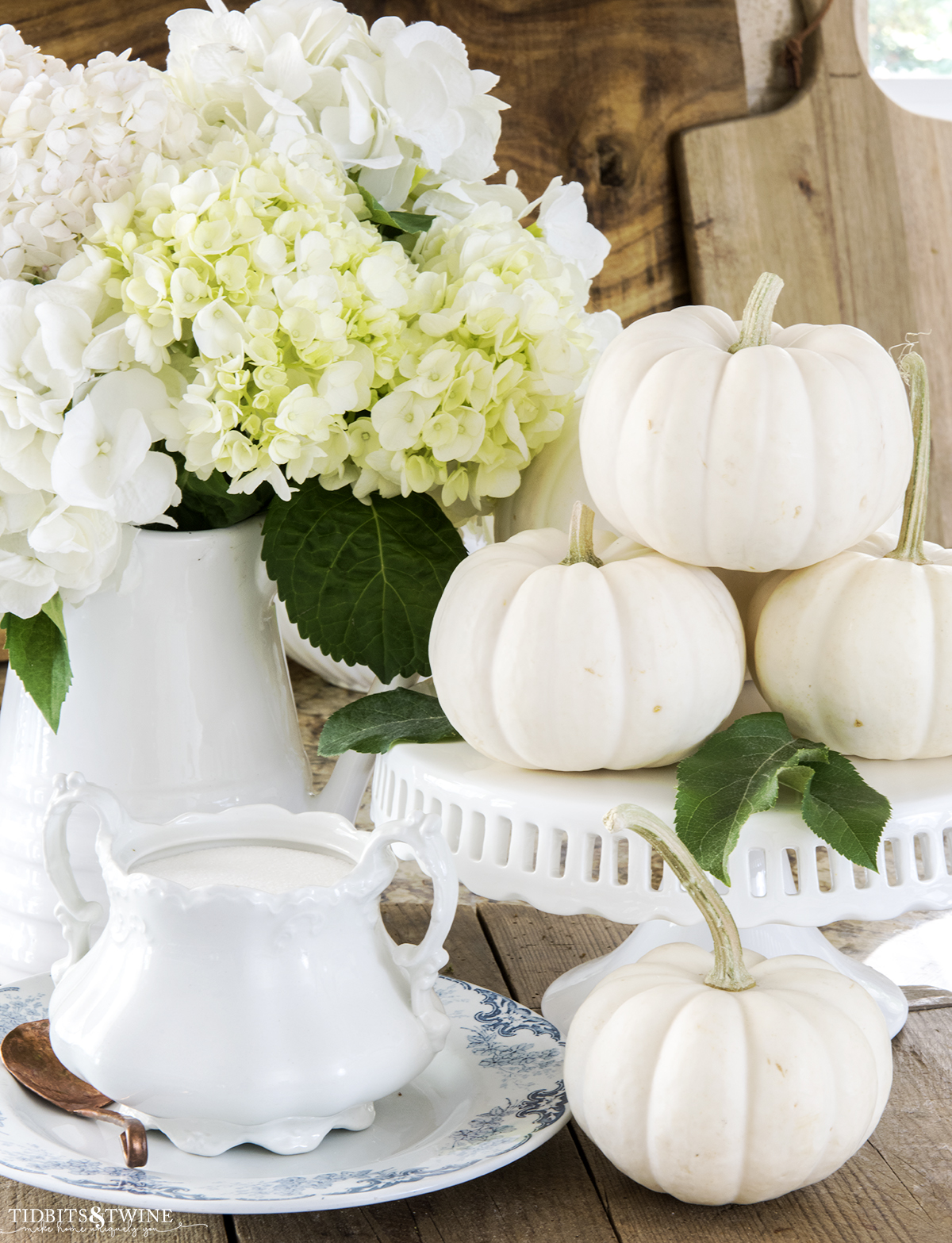 vignette with white pumpkins on a cake stand and white pitcher with white hydrangea and transferware with ironstone sugar bowl