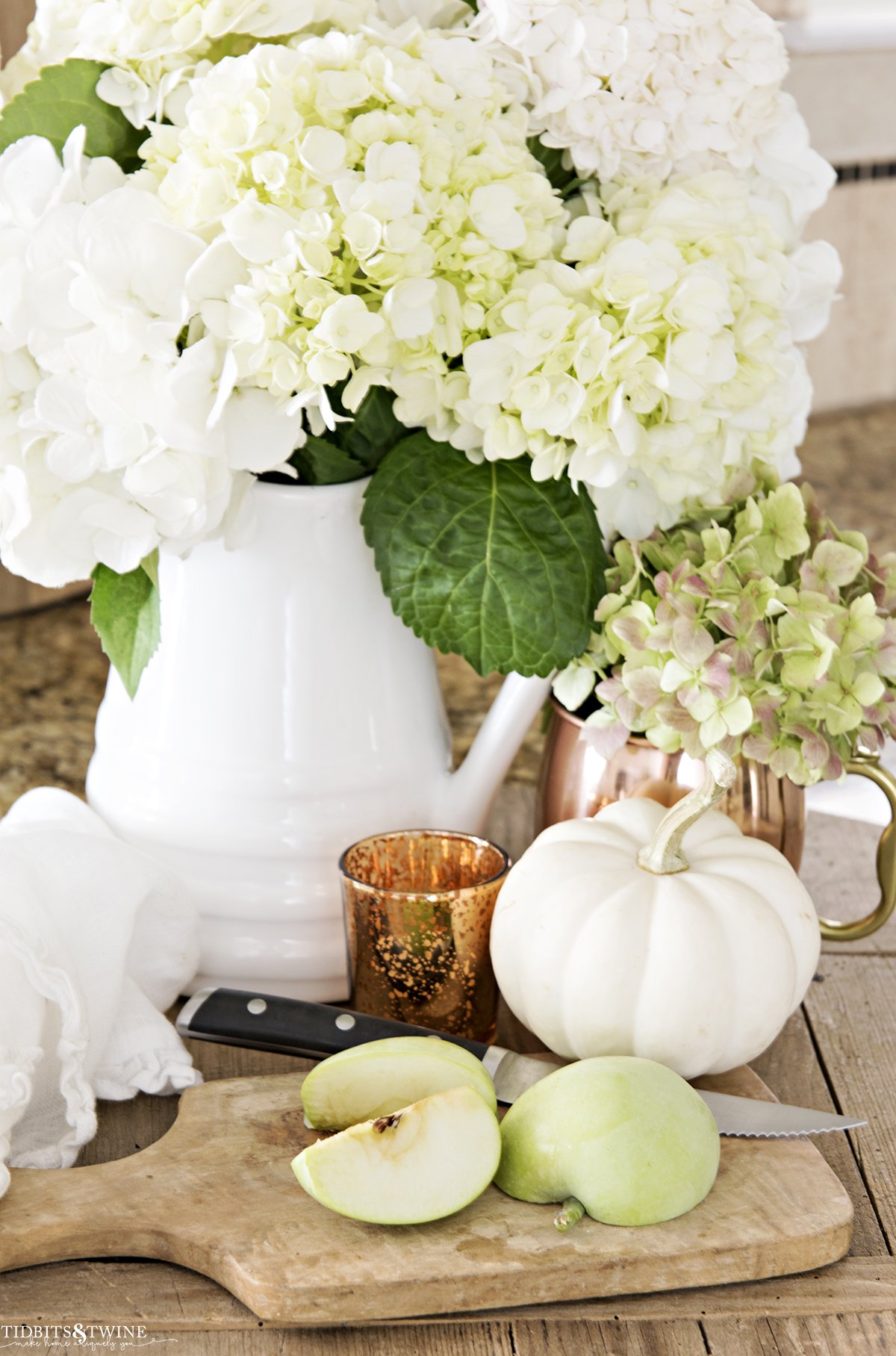 white pitcher with white hydrangeas and copper mug with green hydrangea and cutting board with cut apple