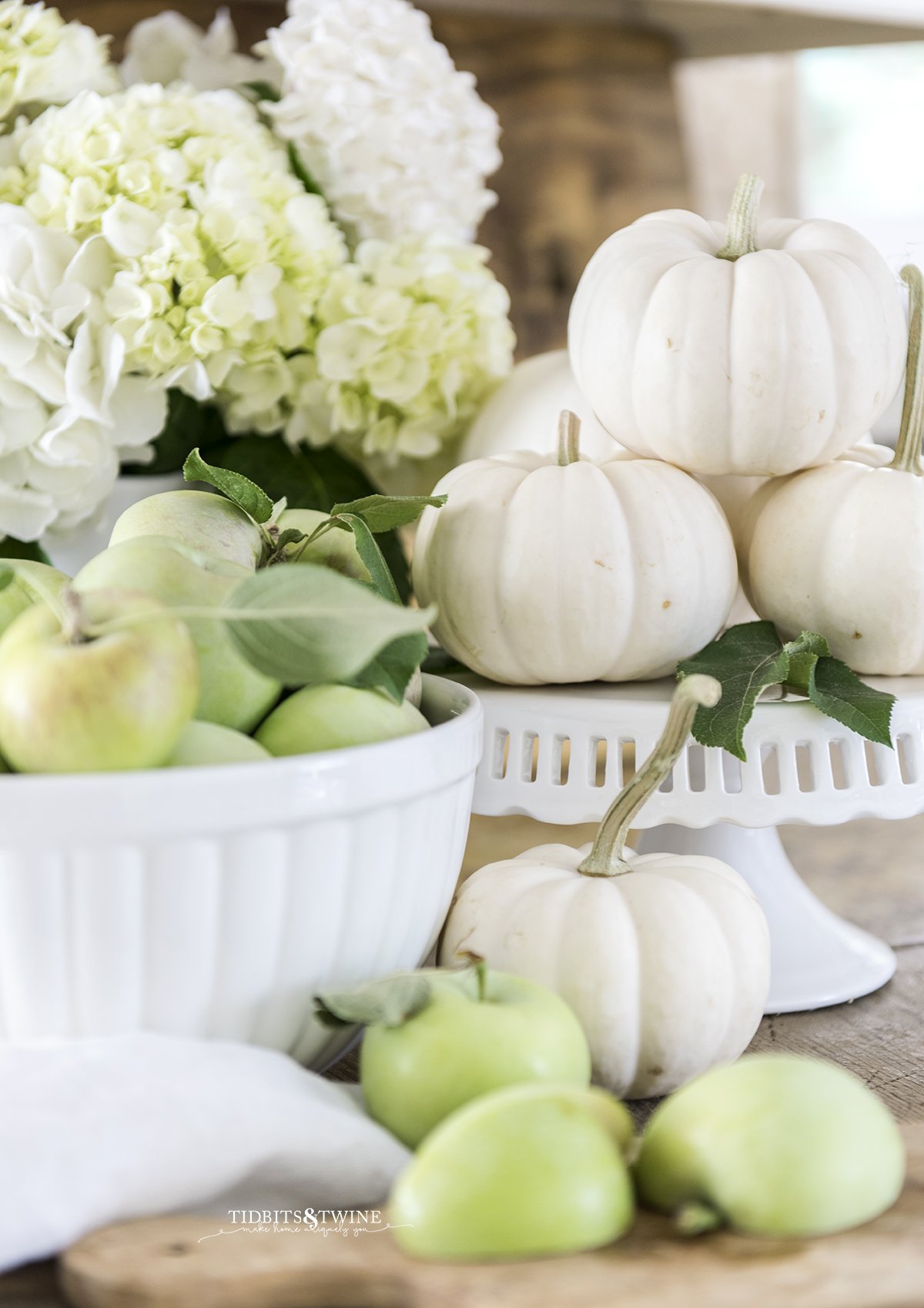 fall kitchen vignette of white bowl full of green apples with white pumpkins on a cake stand and white hydrangea