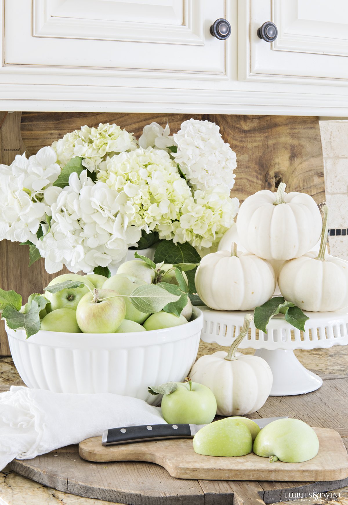 fall kitchen vignette of white bowl full of green apples with white pumpkins on a cake stand and white hydrangea