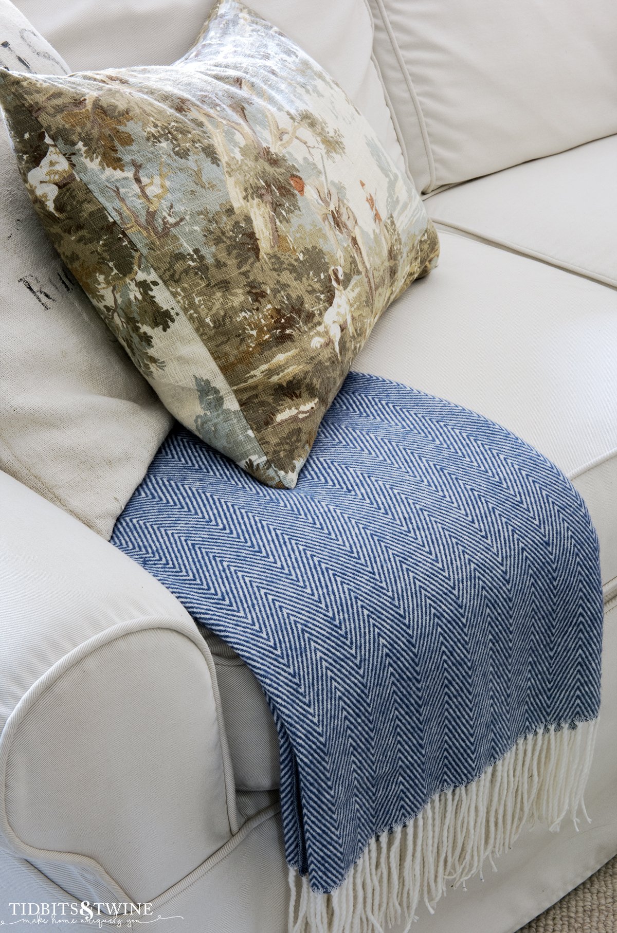 closeup of blue and white herringbone throw with white fringe folded on sofa with fall decorative pillow and grain sack pillow on top