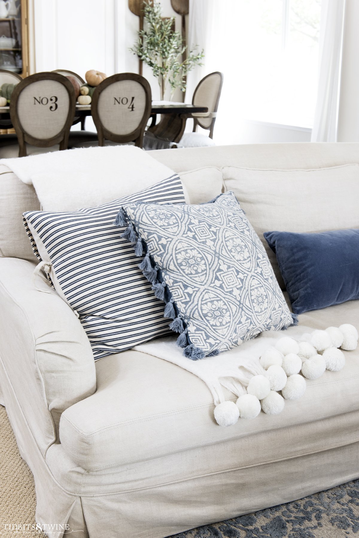Living room sofa with white wool pom throw folded and blue decorative pillows on top and dining room in background