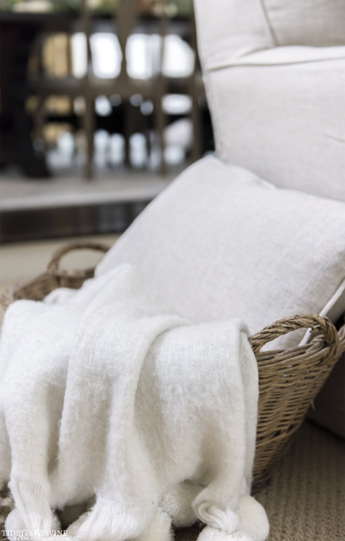 closeup of antique french basket holding a beige pillow and white pom throw on the side of sofa