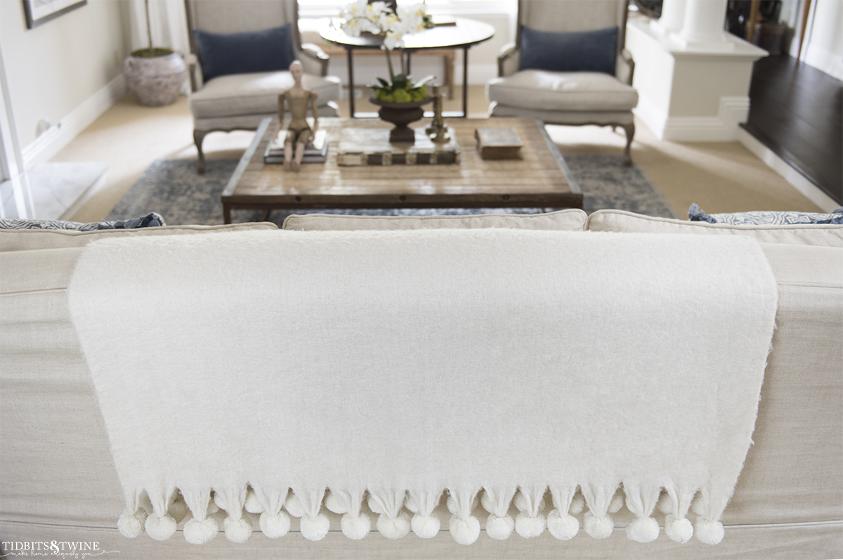 white wool throw with pom fringe hanging down the back of slipcovered sofa in living room with coffee table and chairs in background