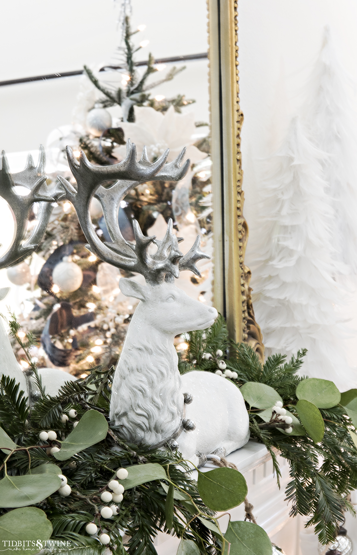 white reindeer with silver antlers on mantel with greenery and gold french mirror behind reflecting a christmas tree