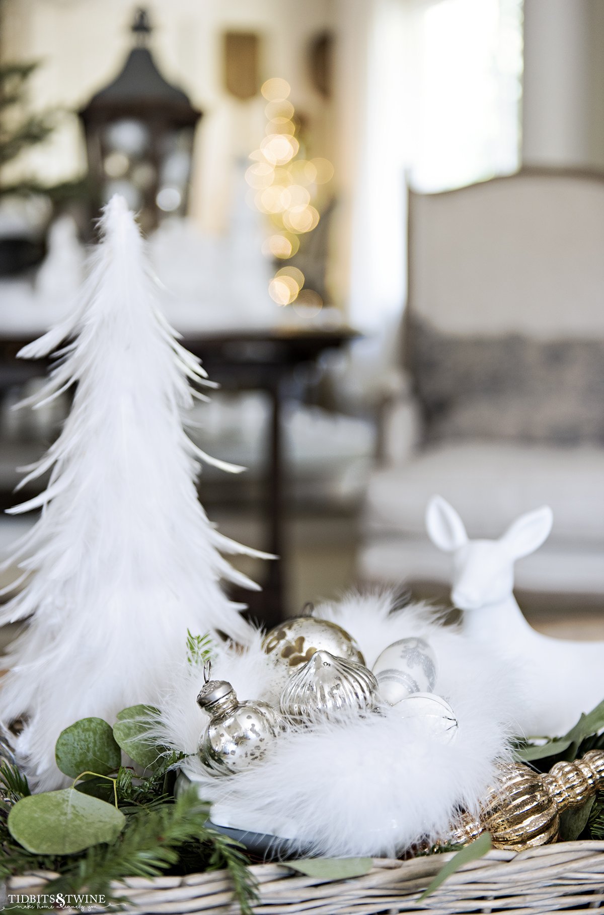 basket tray full of silver ornaments with greenery and white feather tree with seating area in background