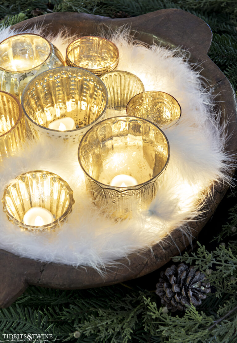 dough bowl full of glass votives with candles and a feather boa tucked around them with Christmas greens around bowl
