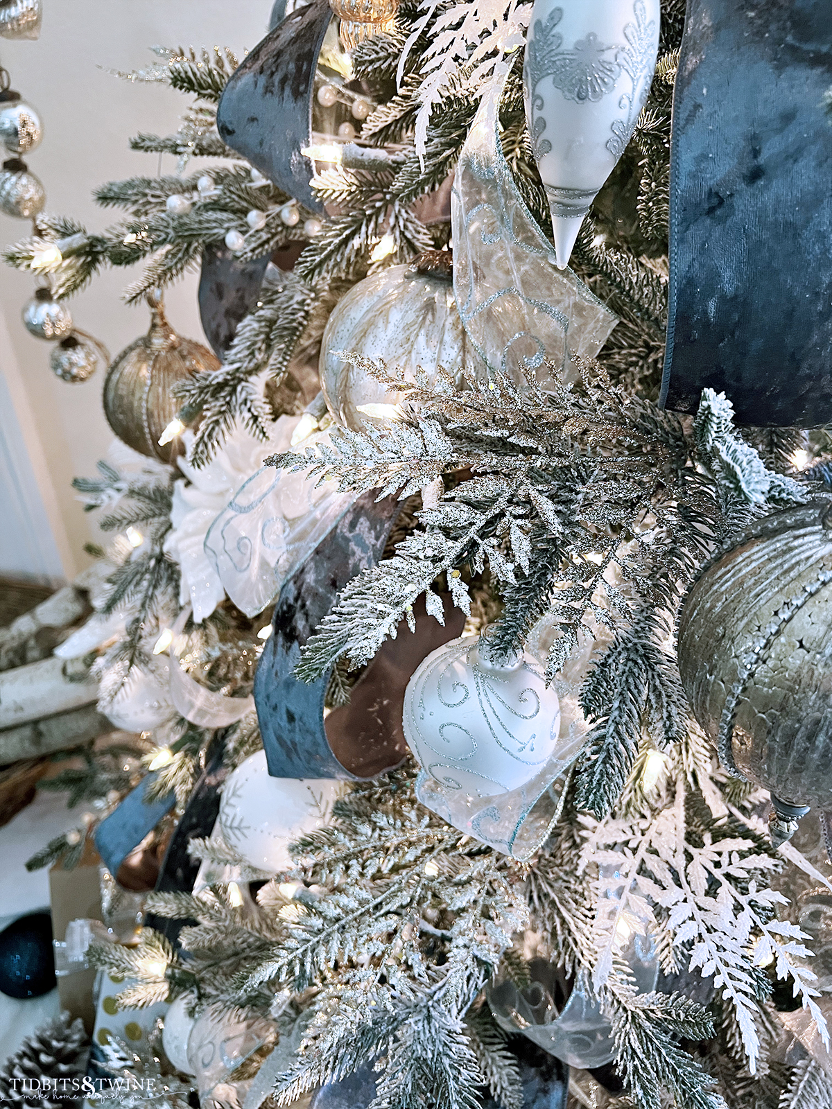 closeup of glitter ferns laying on branches of christmas tree decorated with gold white and silver ornaments and blue ribbon