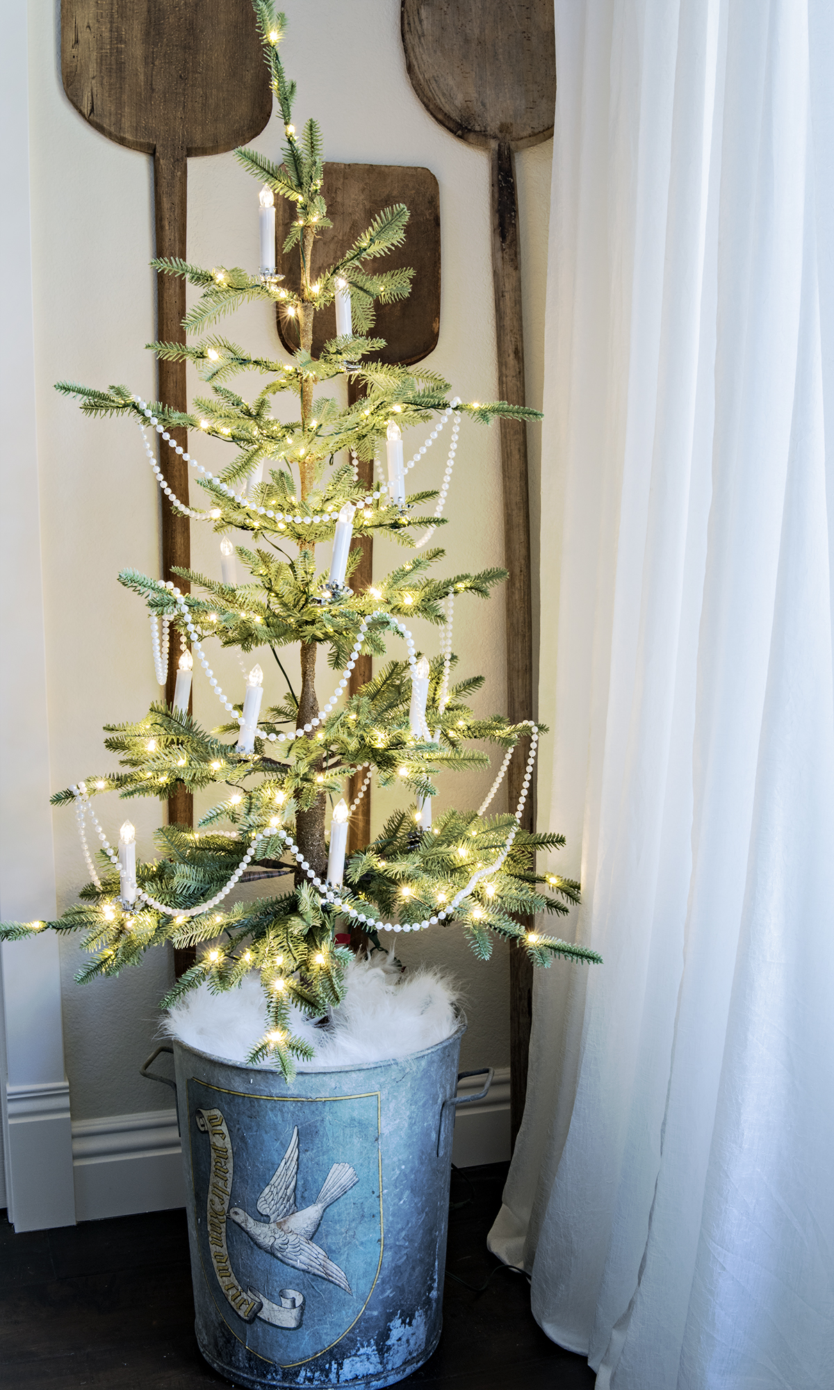 small sparse christmas tree in corner in an antique european bucket with white clip on candles and vintage bread paddles on the wall