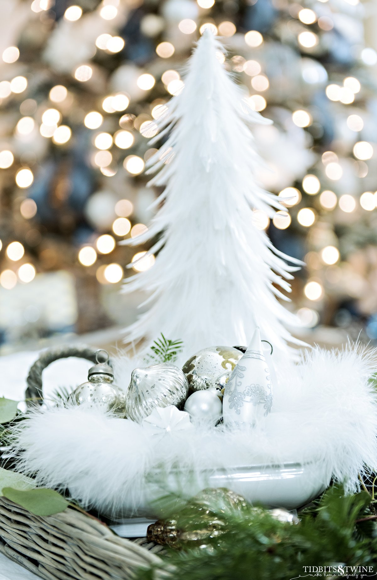 silver ornaments in white ironstone bowl with greenery around and feather tree in background