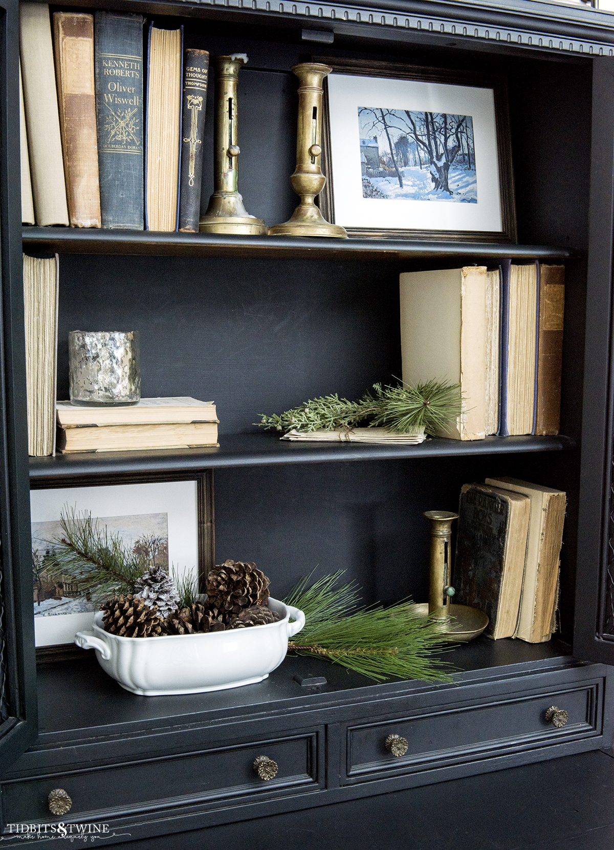 black cabinet with shelves styled for winter decor with vintage books brass candle sticks and evergreens