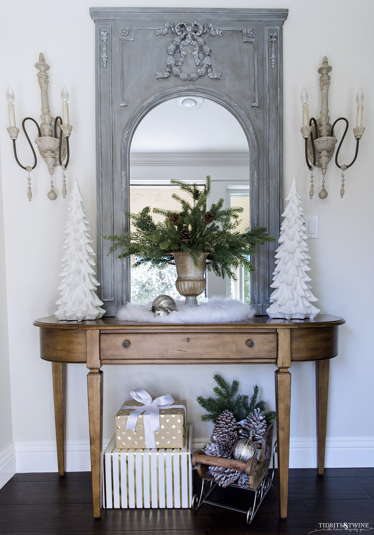 Entryway with small table decorated for Christmas with greens in a french urn flanked by white christmas trees and a blue french trumeau mirror on the wall