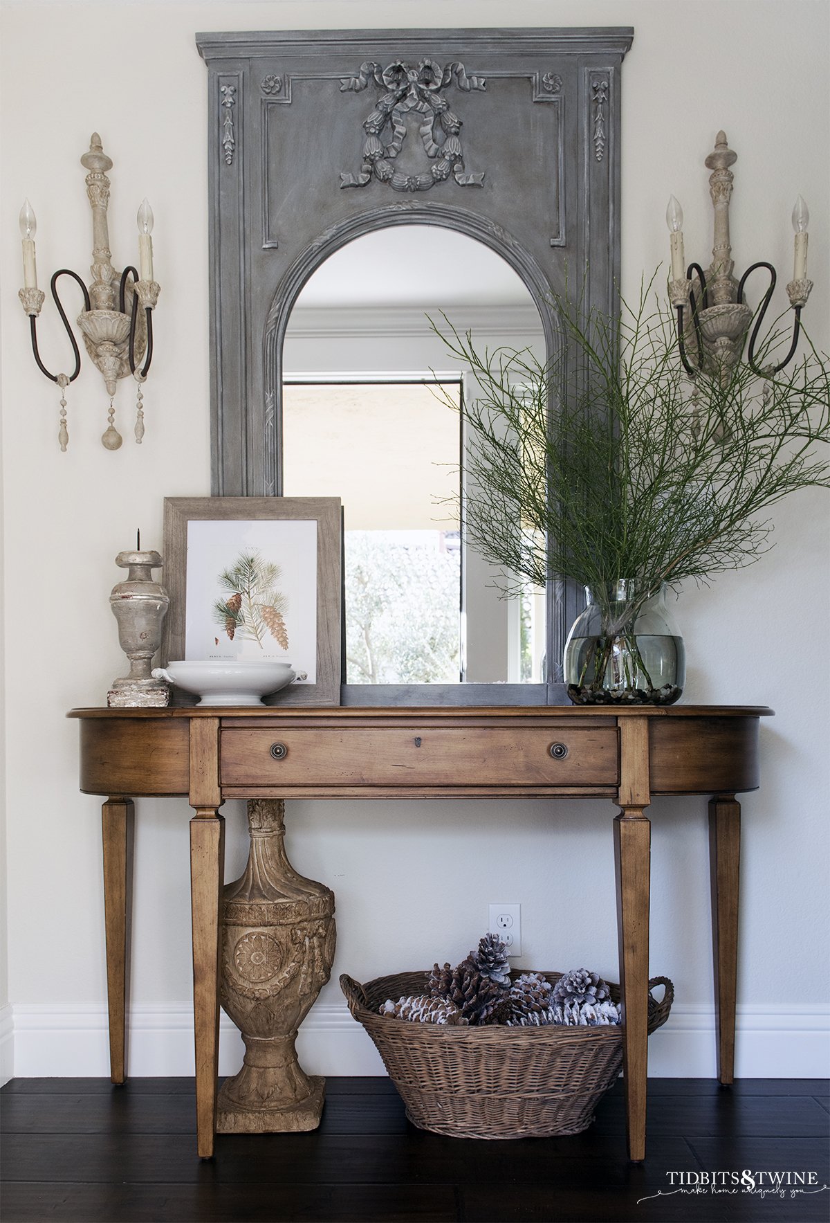 entryway with french trumeau mirror flanked by sconces and small table with winter art and green branches