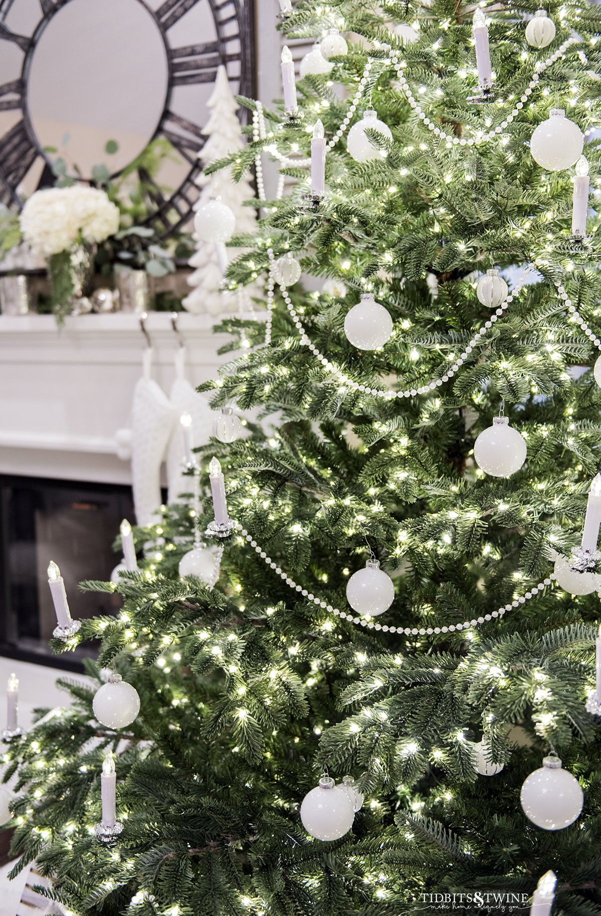 closeup of christmas tree decorated with white ornaments pearl garland and clip on LED candles with fireplace in background