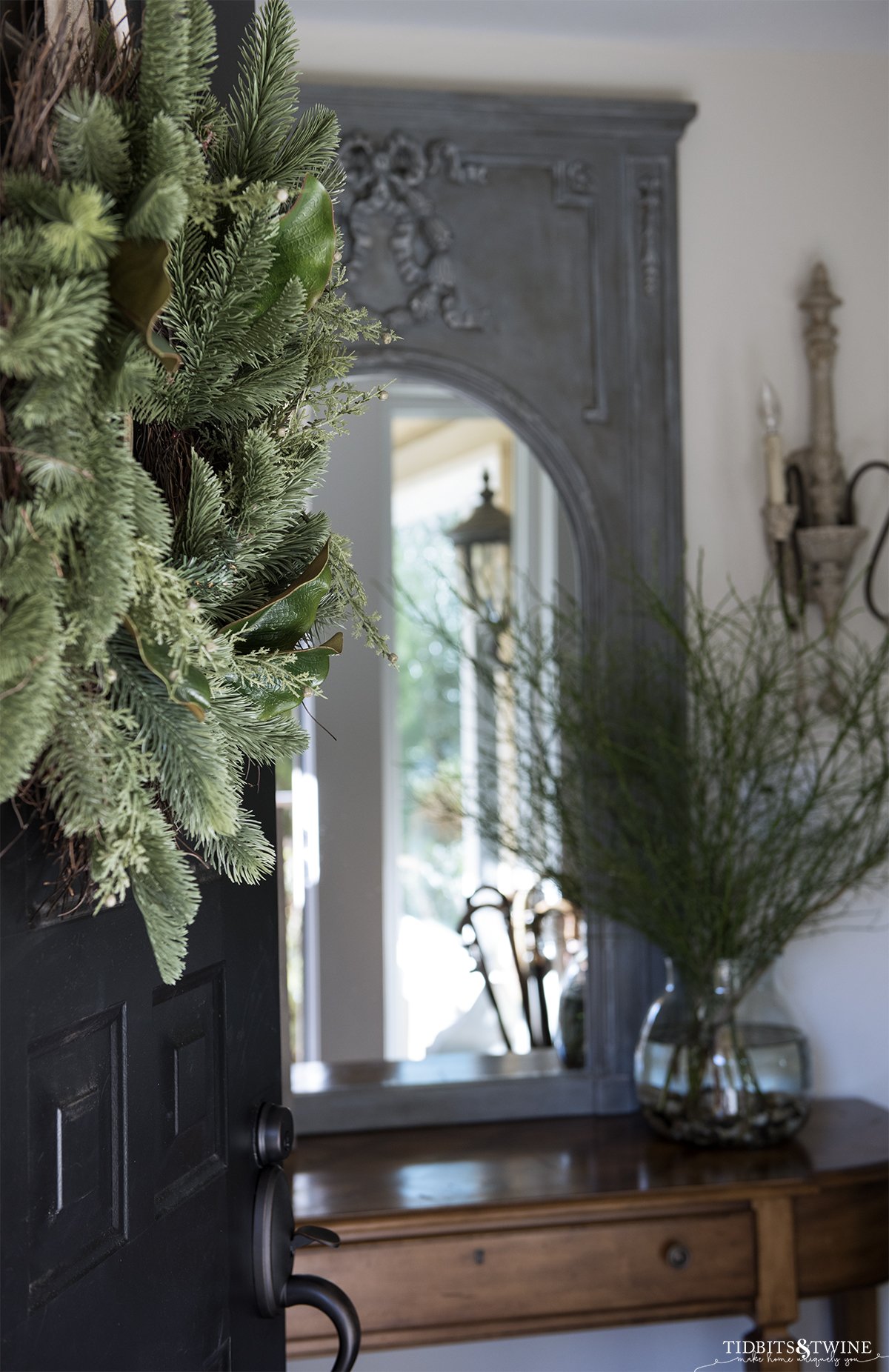 black front door with evergreen wreath partially open to entryway decorated for winter with trumeau mirror