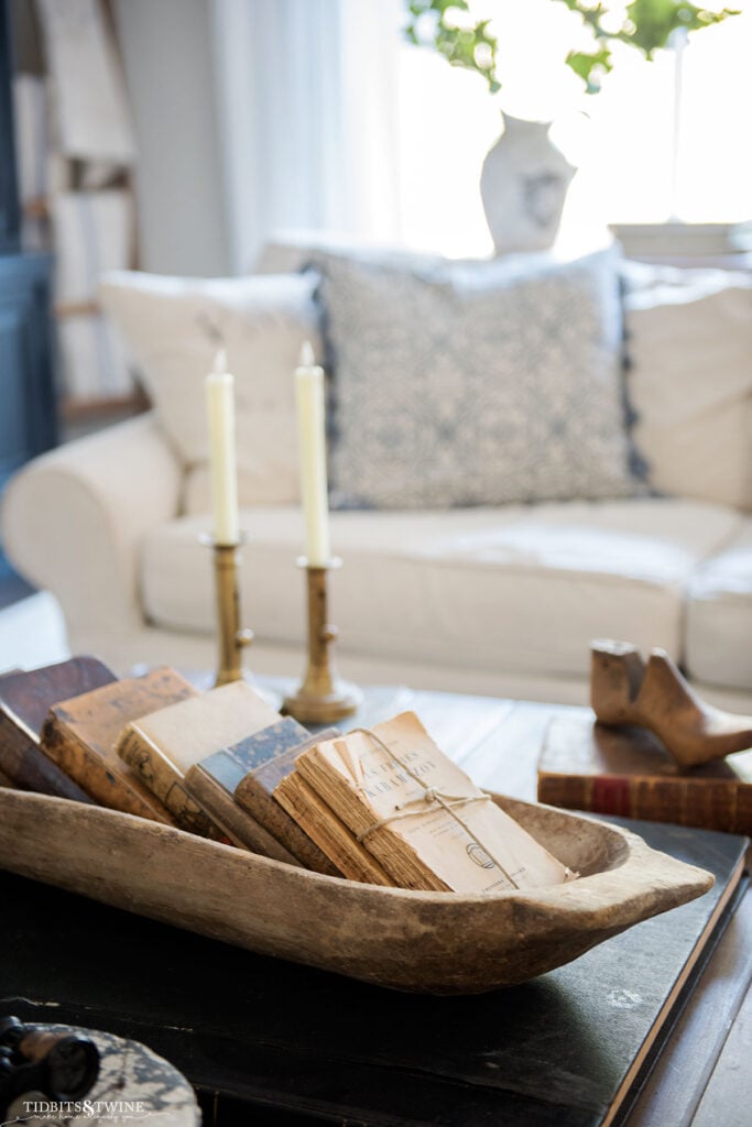 french coffee table styled with antique dough bowl full of old books with two brass candlesticks in background