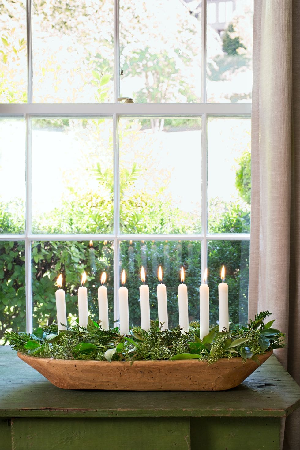 white taper candles in a dough bowl surrounded by greenery on a green console table in front of window