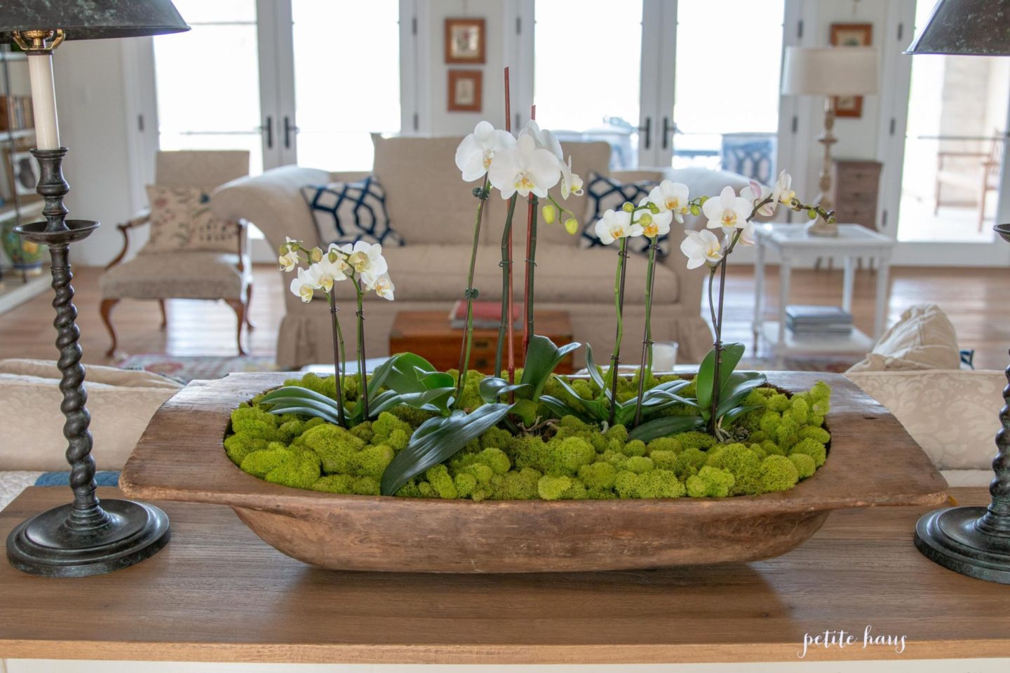 large dough bowl with four white orchids and green reindeer moss on table in living room