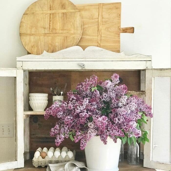 white cabinet with huge display of lilacs in front and two antique bread boards on top